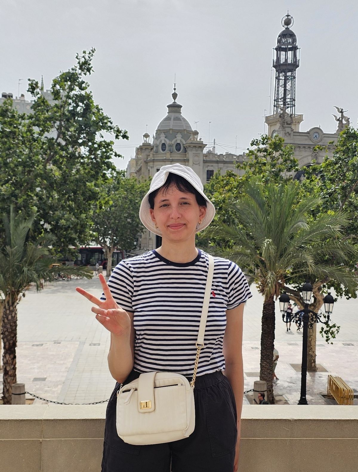 Hanaa smiling to camera in a white bucket hat and a navy-striped t-shirt, standing in a sunlit, tree-lined city square.